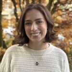 Jenna Sposato, with shoulder-length brown hair, smiles while standing outside. She is wearing a textured white sweater, and the backdrop showcases a tree adorned with autumn leaves in vibrant shades of orange and brown.