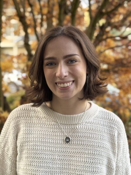 Jenna Sposato, with shoulder-length brown hair, smiles while standing outside. She is wearing a textured white sweater, and the backdrop showcases a tree adorned with autumn leaves in vibrant shades of orange and brown.