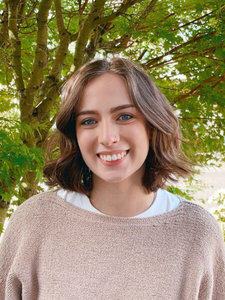 Jenna wearing a beige sweater and smiling in front of a tree with bright green leaves