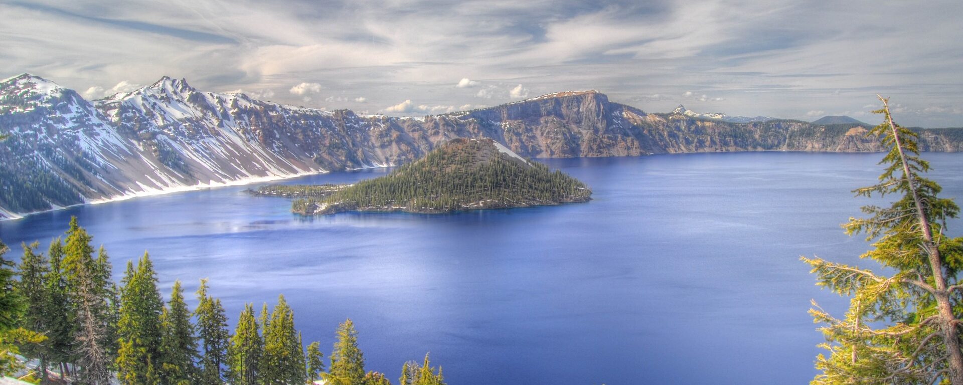 Crater Lake, Oregon. Periwinkle blue water with snow-free trees and ground surrounding the lake.