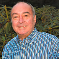 A man in a striped blue shirt smiles at the camera, standing in front of lush green foliage.