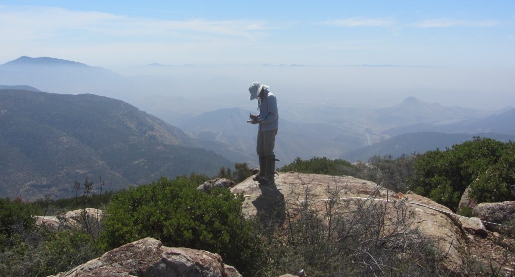 A woman standing on a rock on topo of a mountain taking notes.