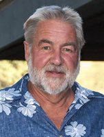 Dr. David Graber wearing a blue Hawaiian shirt with white flowers. Grey hair and beard, tan, dark eyebrows, and a very happy face