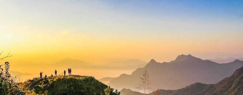 A group of people standing on a hill with a beautiful mountainous view which is both hazy and filled with warm and cool colors