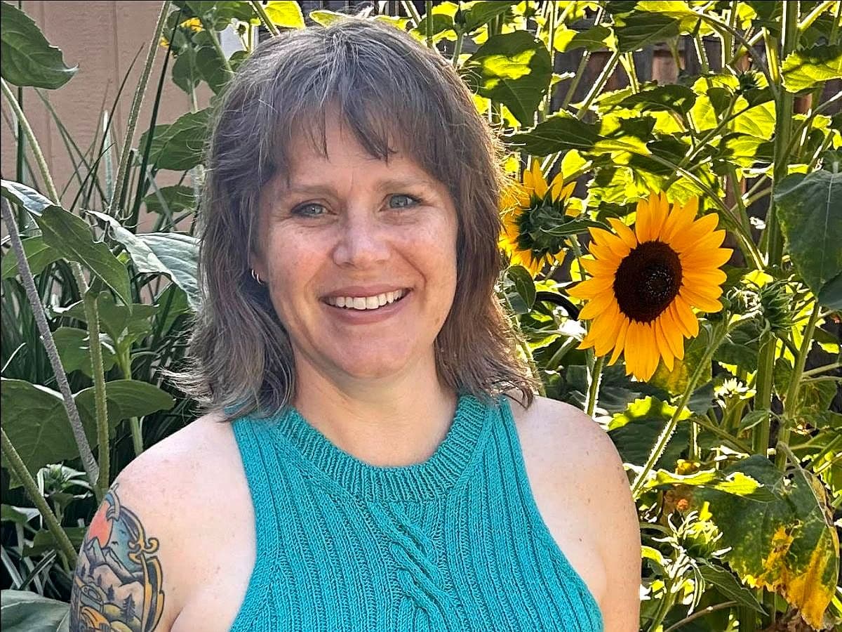 Image of Robin Jones in front of sunflowers wearing a teal-blue high-neck tank top. Robin has short light-brown hair and has a big smile on her face
