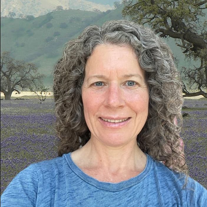Image of Mindy - light-blue crewneck shirt on with grey curly hair and a landscape painting in the background