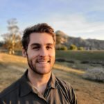 Chris outside at sunset wearing a brown button-down shirt with green and brown grass visible behind him.