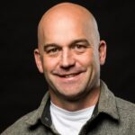 A bald man with a friendly smile, identified as Dr. Chas Jones, is wearing a grey jacket over a white shirt, set against a dark background.