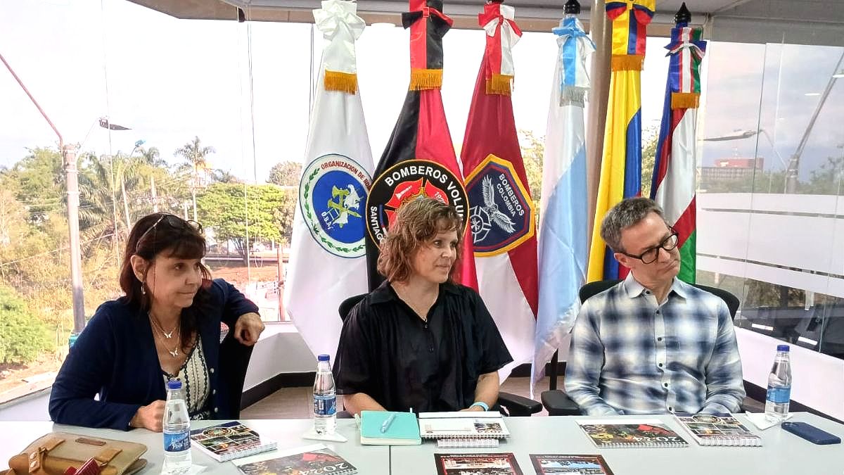 Three people sit at a table with brochures, in front of flags from various countries. The conference room, set for the November 2024 CBI Newsletter meeting, features large windows that flood the space with light.