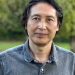 Masami Takikawa, a software developer with dark hair, stands outdoors in a denim shirt, framed by lush greenery in the background.