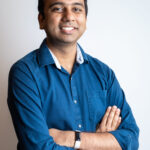 Wearing a blue shirt and a smile, Samapriya Roy stands confidently with arms crossed against a plain white background, embodying the spirit of the awesome Google Earth Engine Community Catalog.