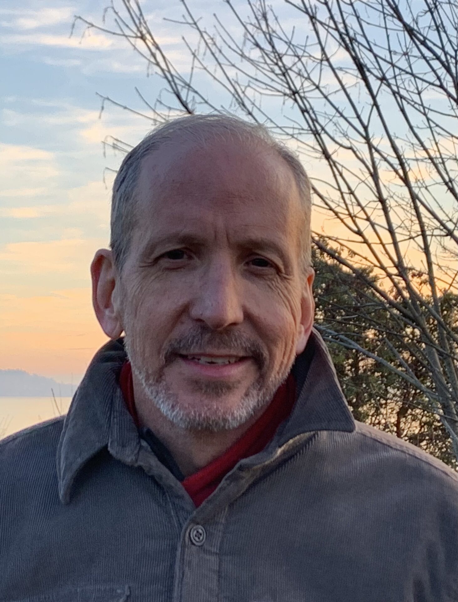 Man smiling outdoors with a sunset sky in the background. Hes wearing a button-up jacket, and there are leafless tree branches to the side.