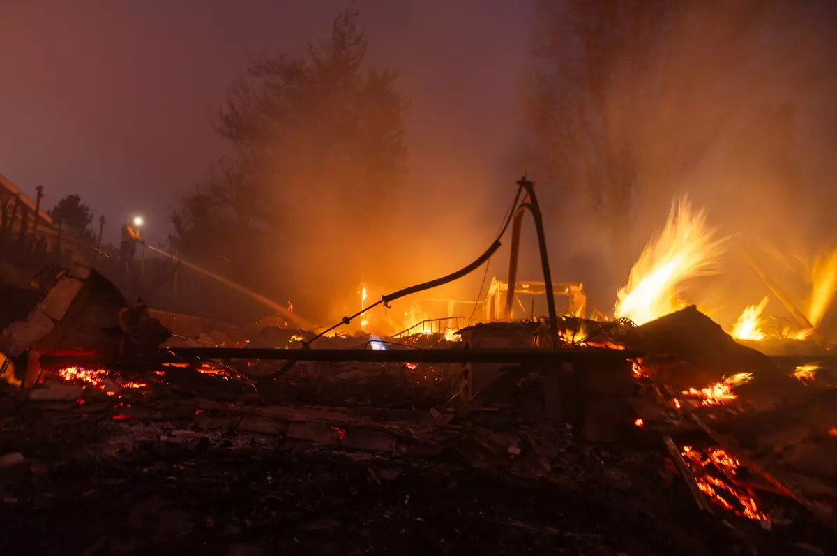 A landscape engulfed in flames showing a fire scene at night. Firefighters work to control the blaze, with smoke rising into the night sky. The silhouette of trees is visible against the orange and red glow of the fire.