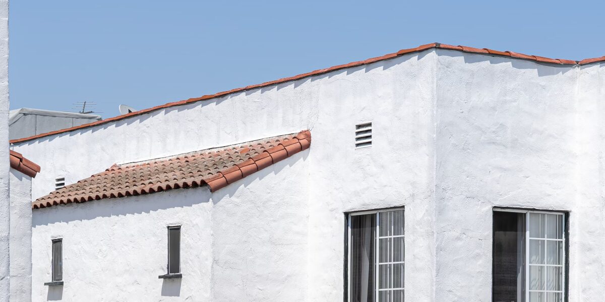 White stucco building with red-tiled roof sections against a clear blue sky. The building features a few narrow windows with vertical bars and a louvered vent near the roofline.