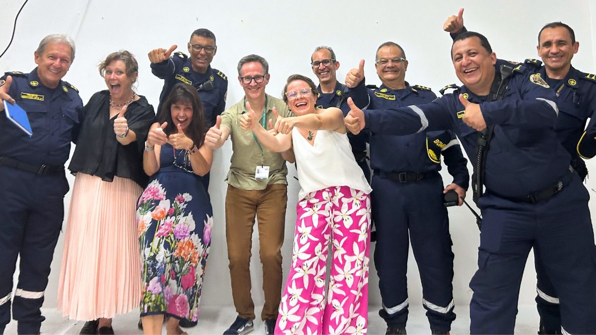 A group of people, including men and women, are standing together, smiling and giving thumbs up. Some wear navy uniforms with gold trim, while others wear casual clothing. They pose happily against a plain white background.