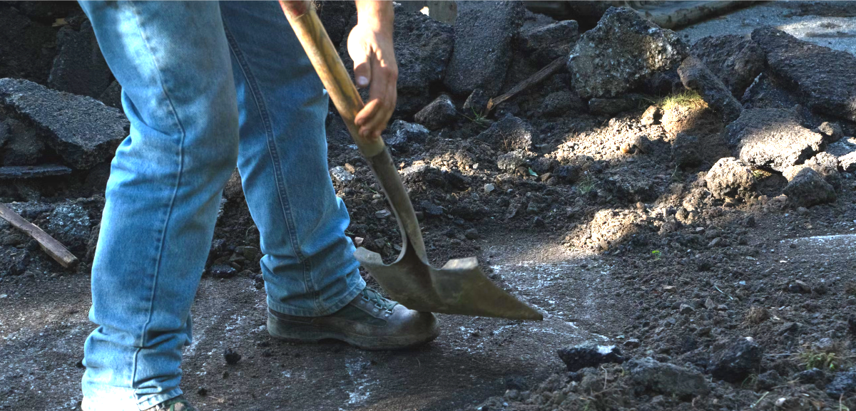 Person in blue jeans and work boots shoveling dirt and rocks on a construction site. Partial view of their legs and hands holding the shovel. Rubble and broken asphalt are visible in the background.