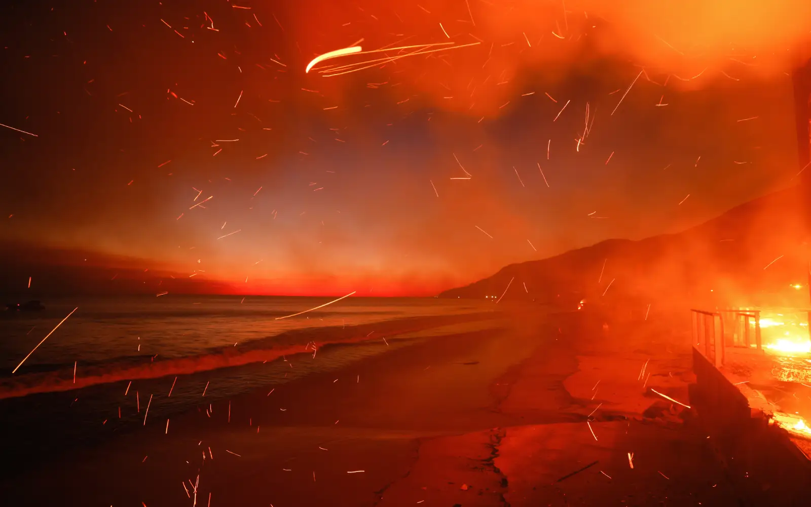 A dramatic scene of a coastline fire at dusk. Sparks and embers fill the air, illuminating the red-orange sky. The ocean contrasts with the fiery glow, and silhouettes of hills are visible in the distance.