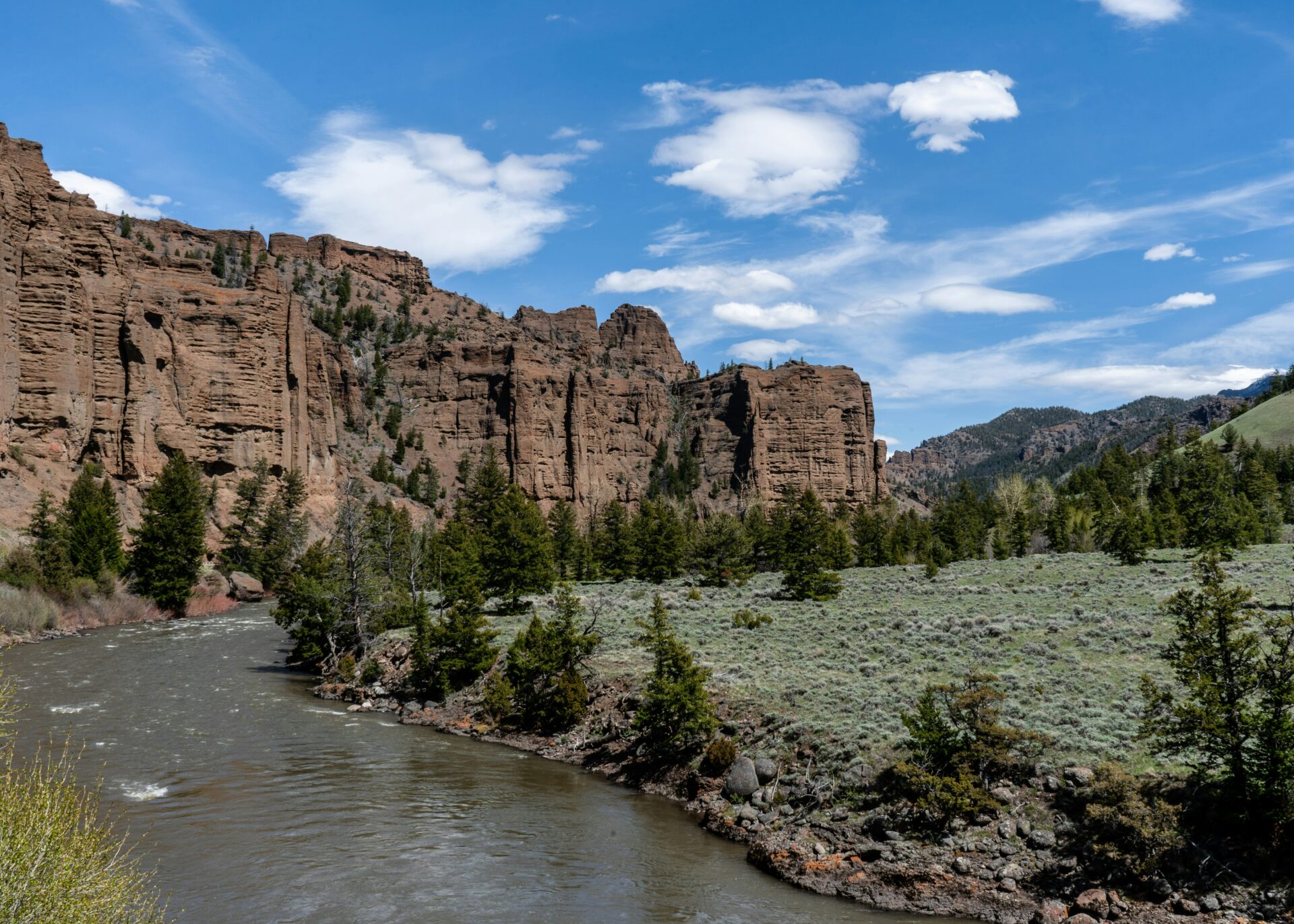 A scenic landscape featuring a river winding through a valley with rugged rock formations and cliffs in the background. Pine trees and grassy fields surround the river under a clear blue sky with a few scattered clouds.