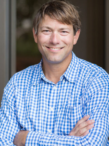 Smiling man in a blue and white checkered shirt stands with arms crossed in an indoor setting with a glass wall in the background.