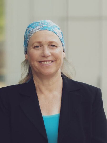 A woman with long hair, wearing a blue patterned headscarf and a black blazer, stands smiling in front of a glass building. Her reflection is visible on the glass. The background is softly blurred.