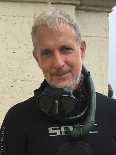 Jeff Tracey wearing a black wetsuit and scuba diving mask stands in front of a stone wall. His snorkel is draped over his shoulder, and the sea is visible in the background.